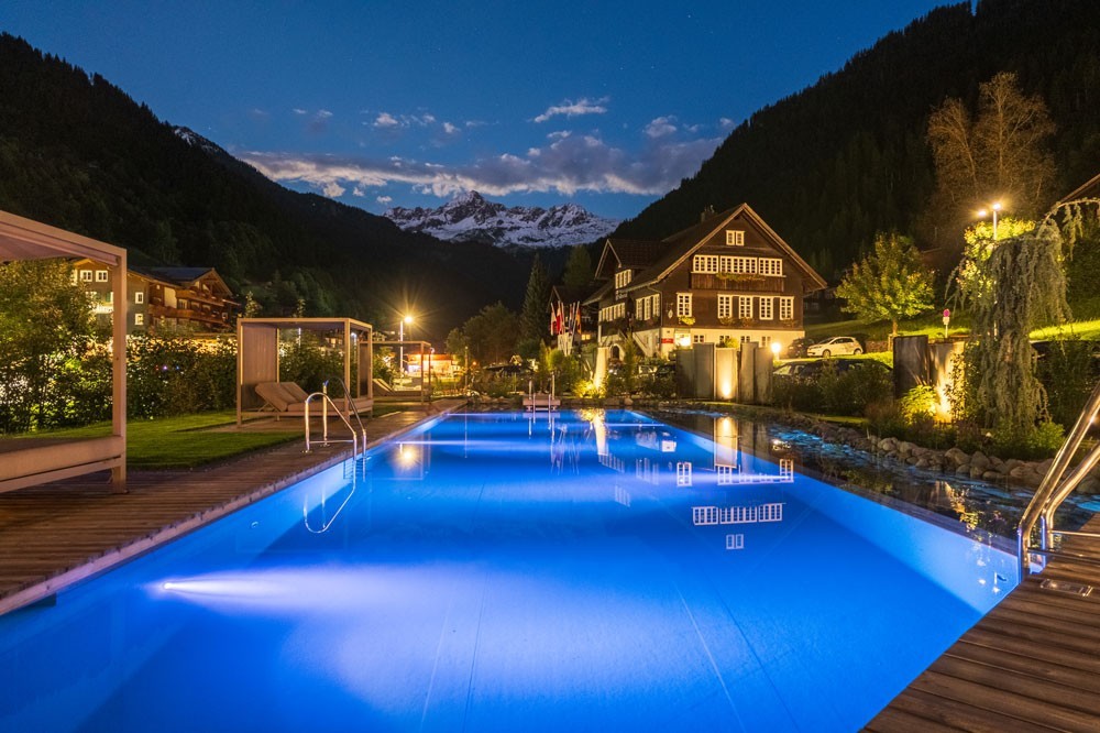 Illuminated hotel pool at night in front of a mountain village and the Alps